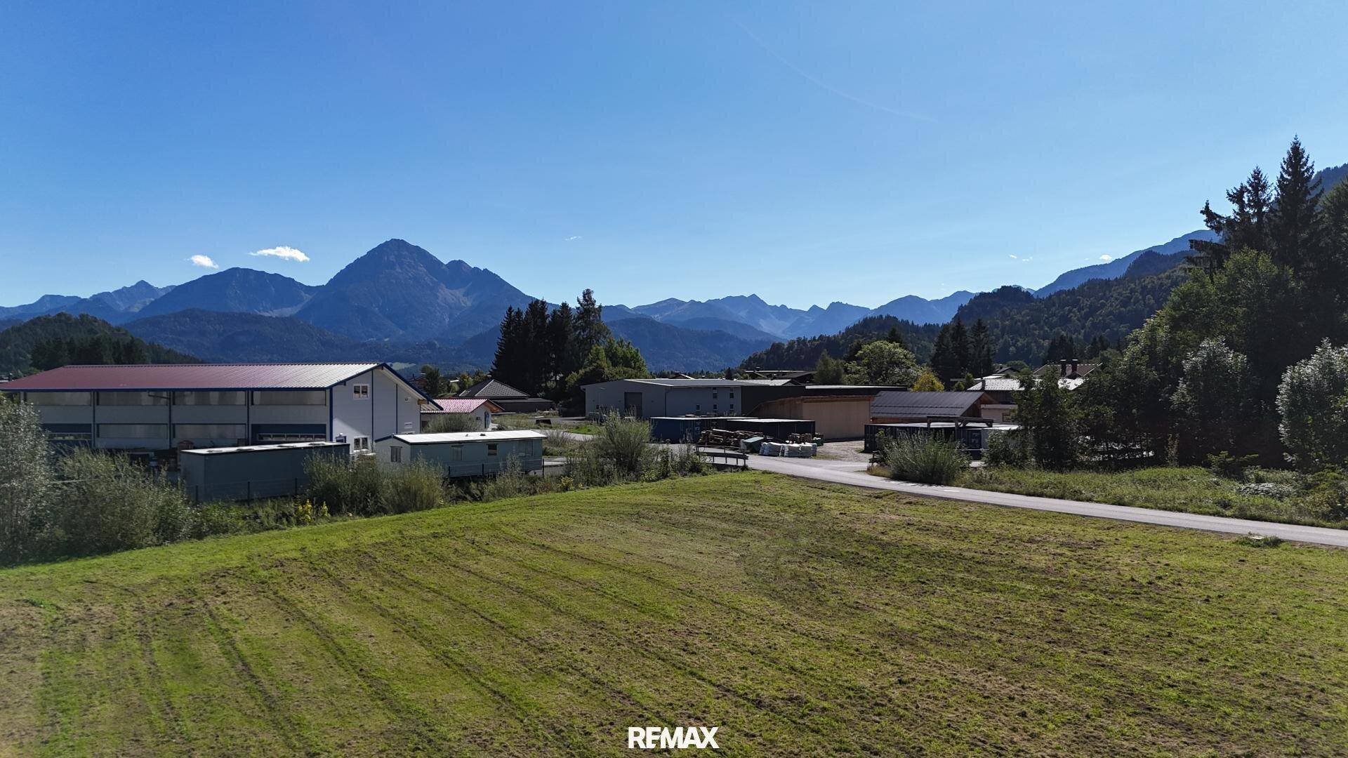 Weitläufige Landschaft mit Feldern, Gebäuden und majestätischen Bergen im Hintergrund unter blauem Himmel.