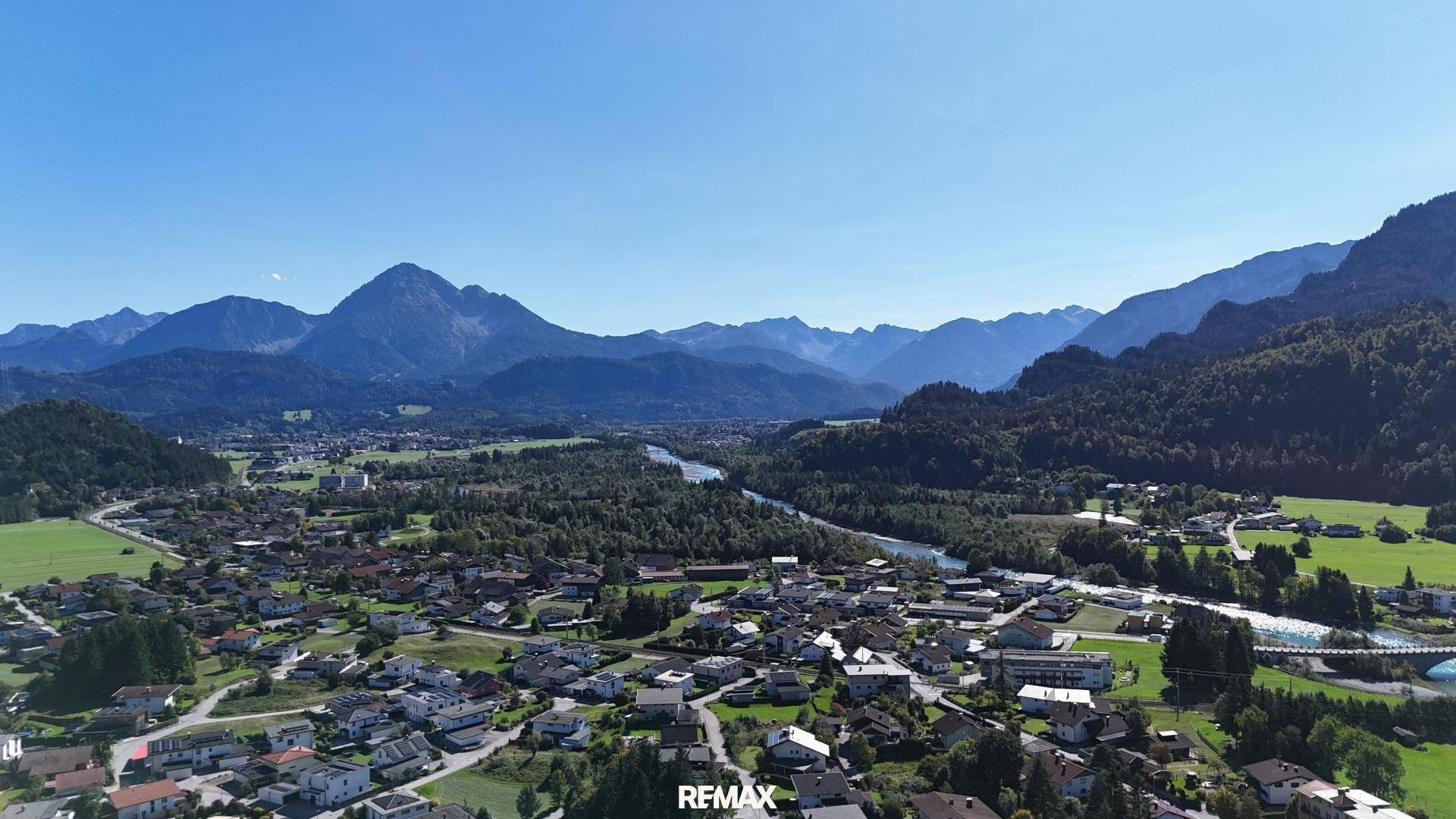 Panoramablick auf eine Siedlung im Tal mit Fluss, Wäldern und beeindruckender Bergkulisse unter klarem Himmel.