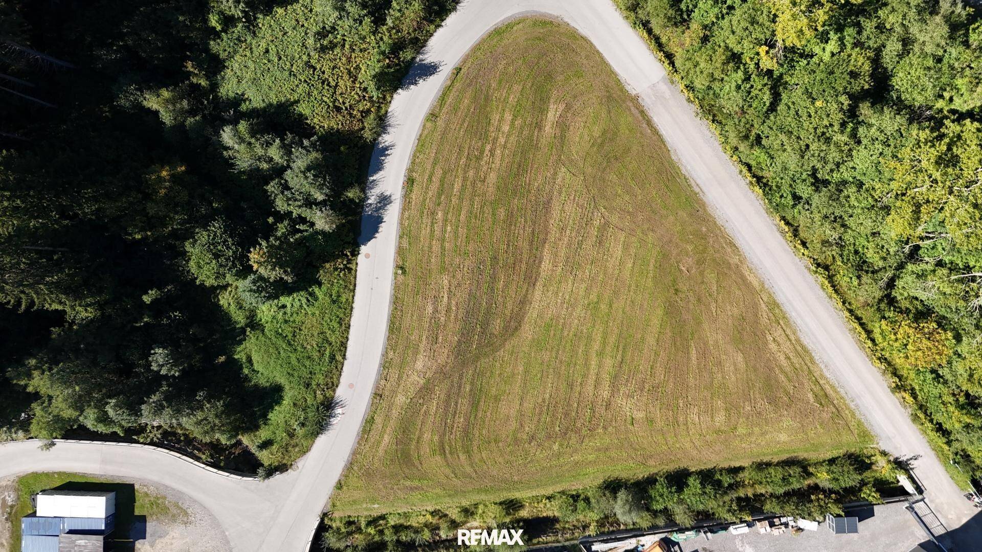 Luftaufnahme eines unbebauten Grundstücks, umgeben von Wald und Zufahrtsstraßen, bei sonnigem Wetter.