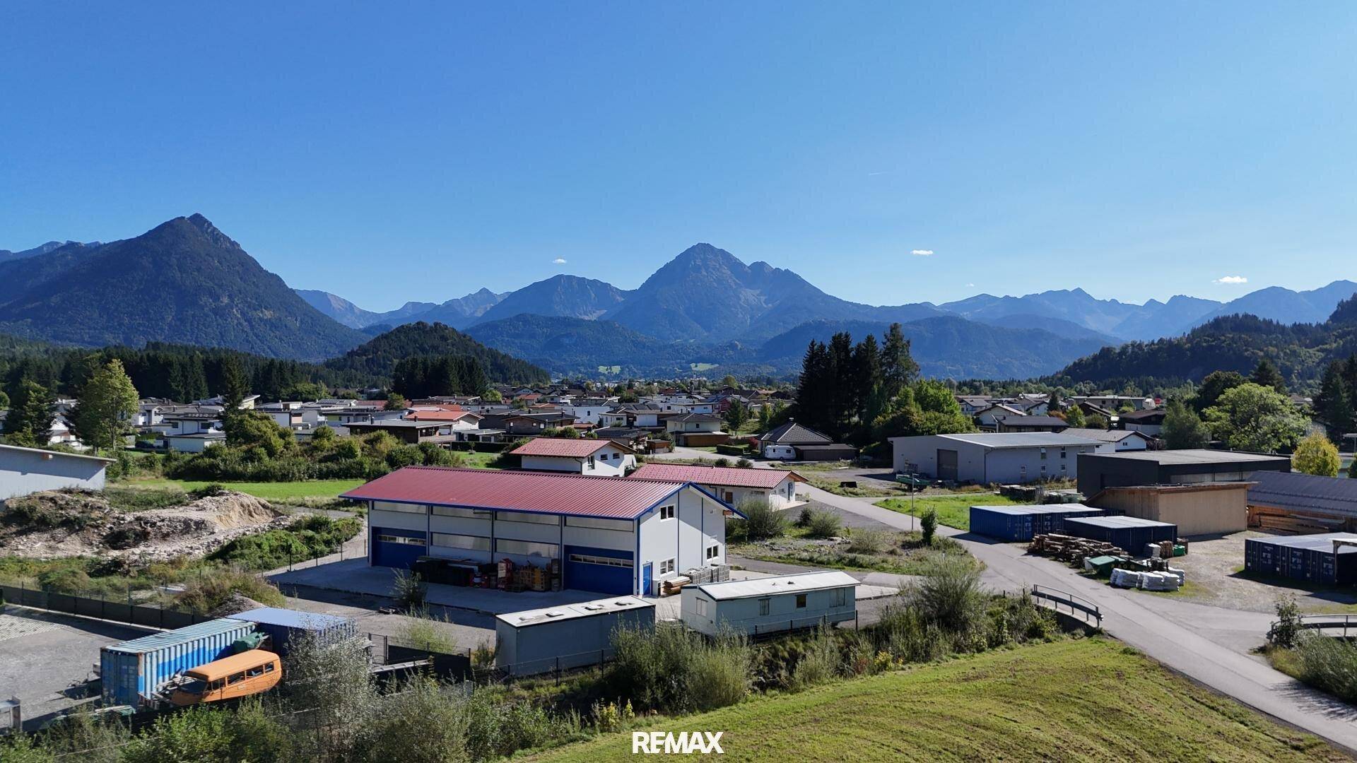 Panoramablick auf eine ländliche Siedlung mit Bergen und weiten Grünflächen unter blauem Himmel.