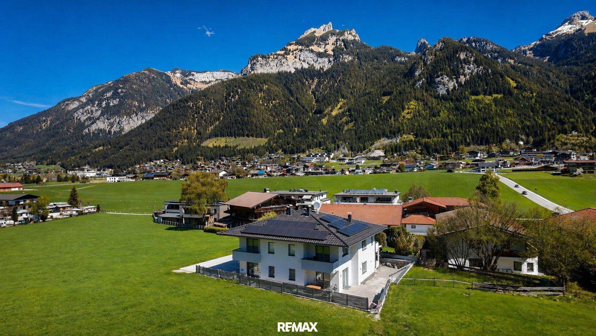 Modernes Haus mit Photovoltaikanlage auf dem Dach, umgeben von weiten grünen Feldern und Bergen.