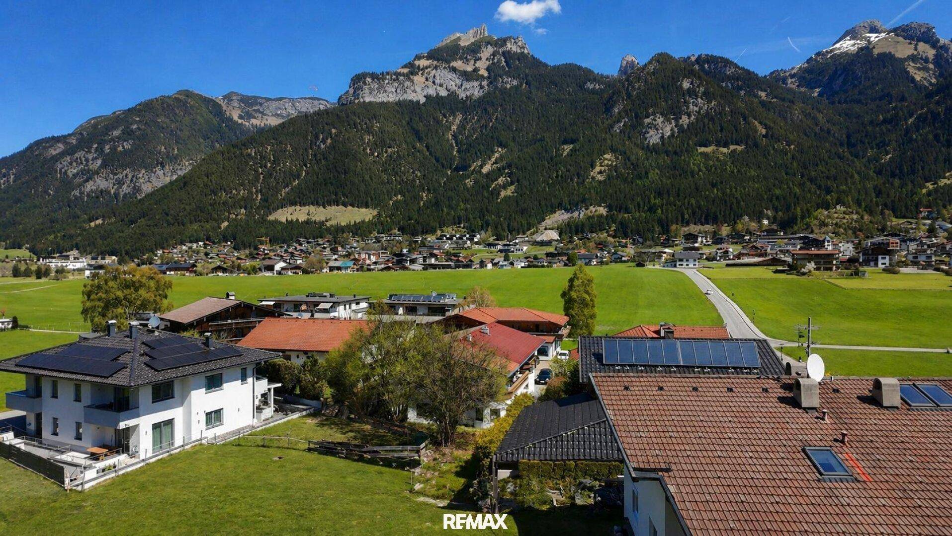 Luftaufnahme von Häusern mit Solaranlagen und traditionellen Dächern, umgeben von grünen Wiesen und Bergen.