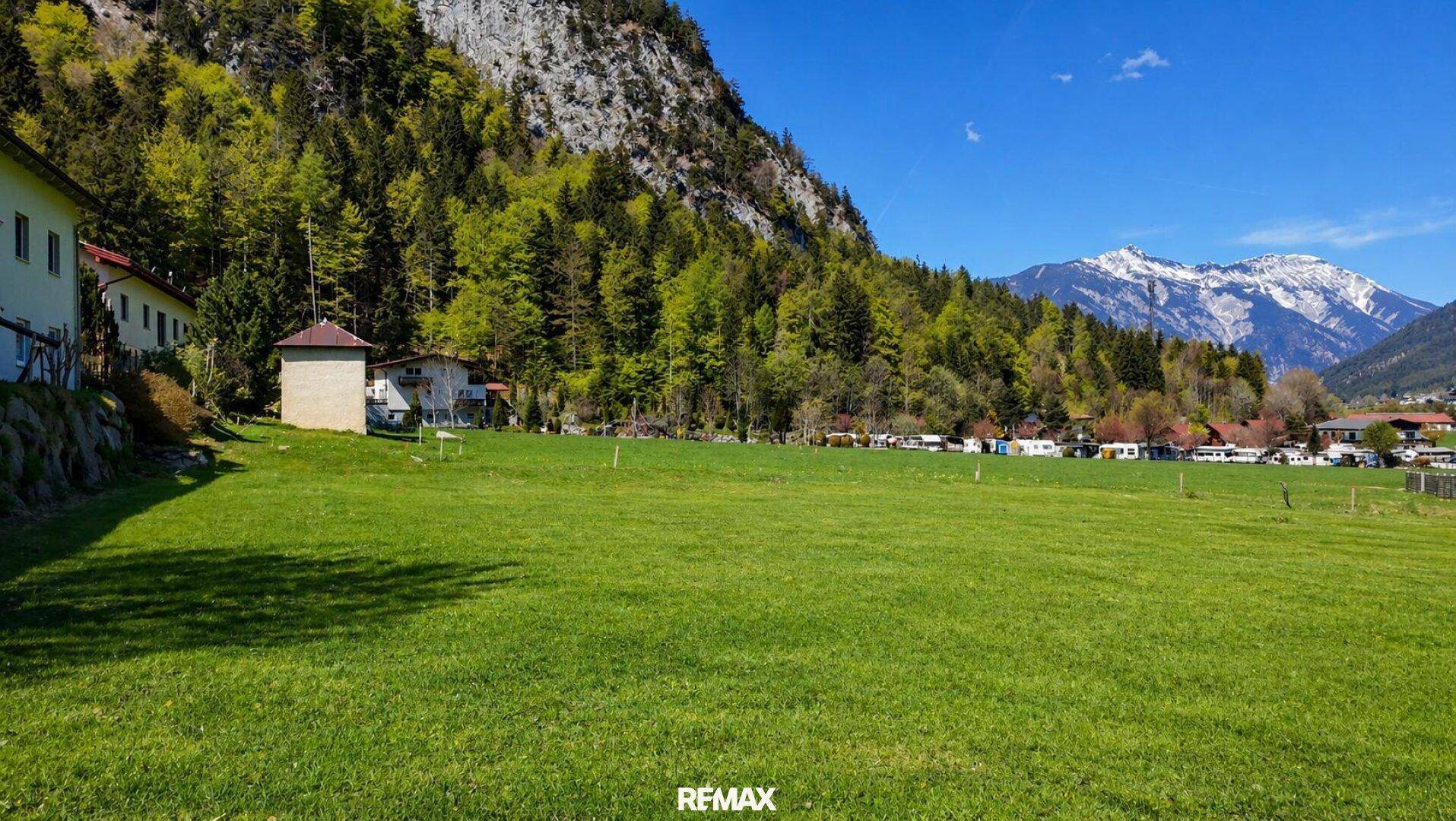 Weite grüne Wiese mit Blick auf bewaldete Berge und einige Häuser in der Ferne.