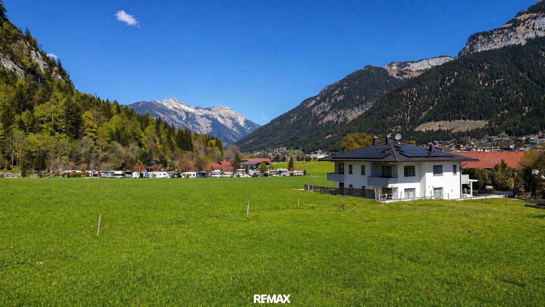 Weite grüne Wiese mit einem modernen Haus und einem Campingplatz im Hintergrund, umgeben von Bergen.