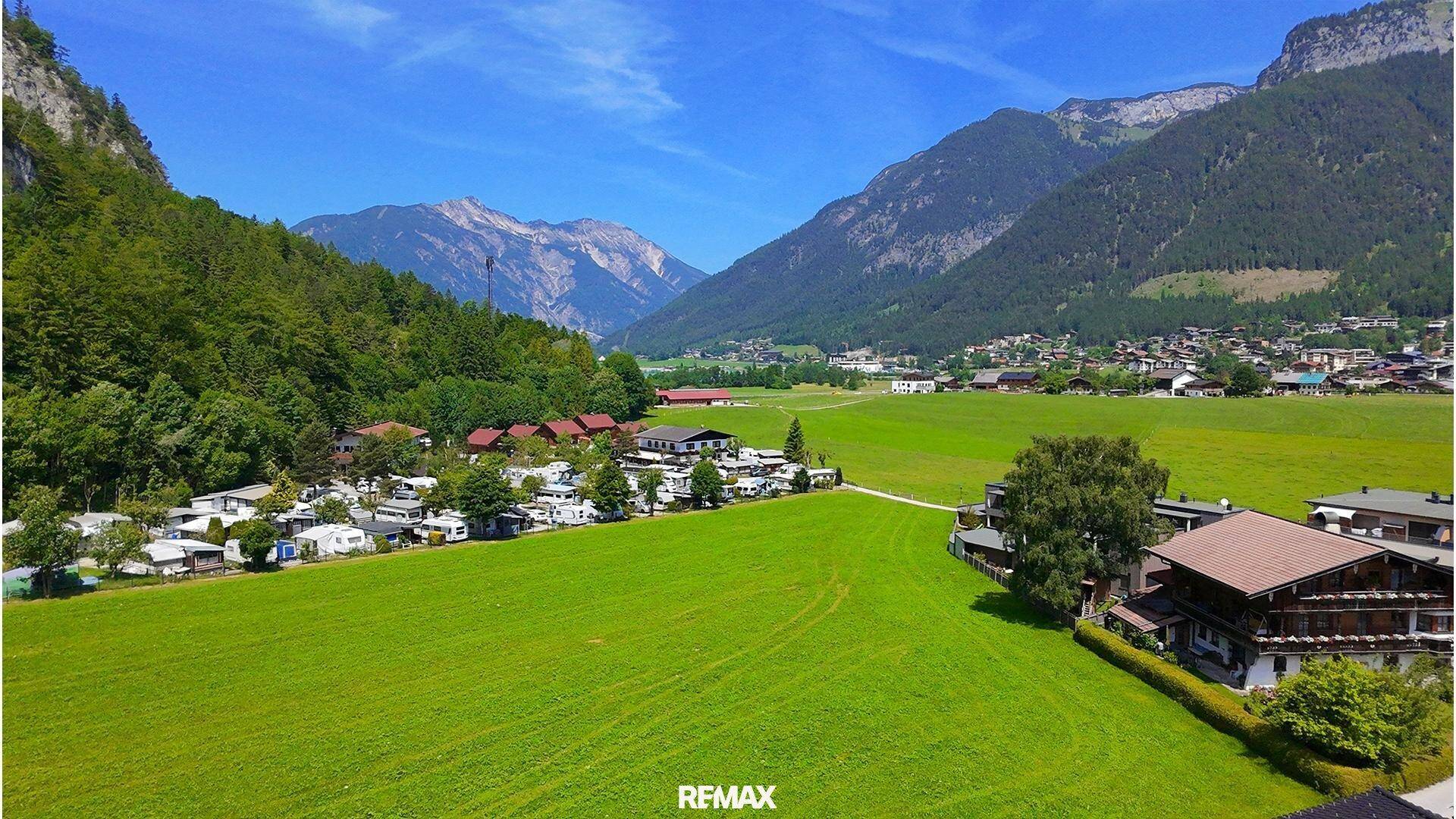 Panoramablick auf ein grünes Tal mit Campingplatz und Dorf, umgeben von majestätischen Bergketten.