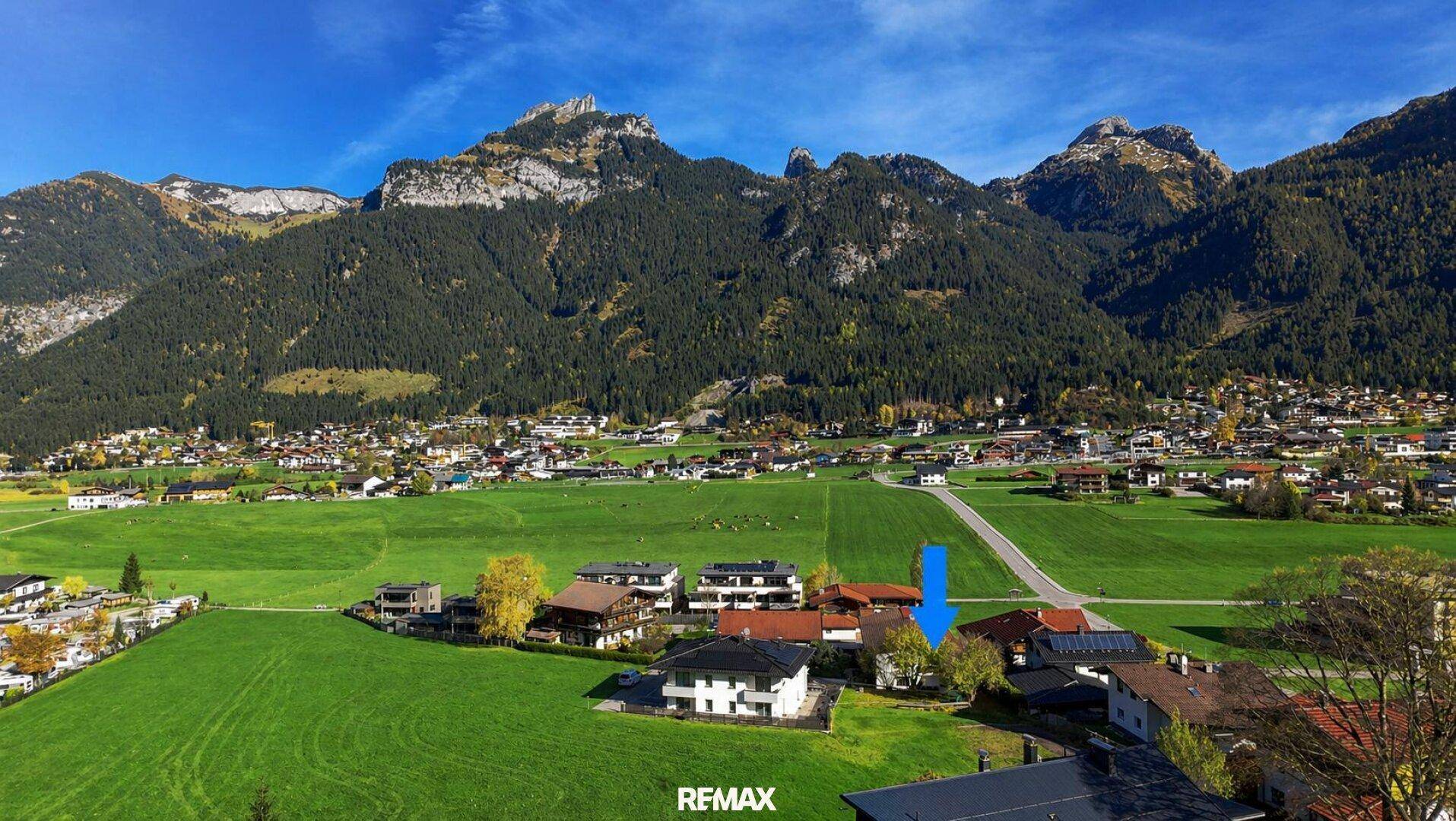 Luftaufnahme eines Dorfes in den Bergen mit grünen Feldern und einem blauen Pfeil, der auf ein Gebiet zeigt.