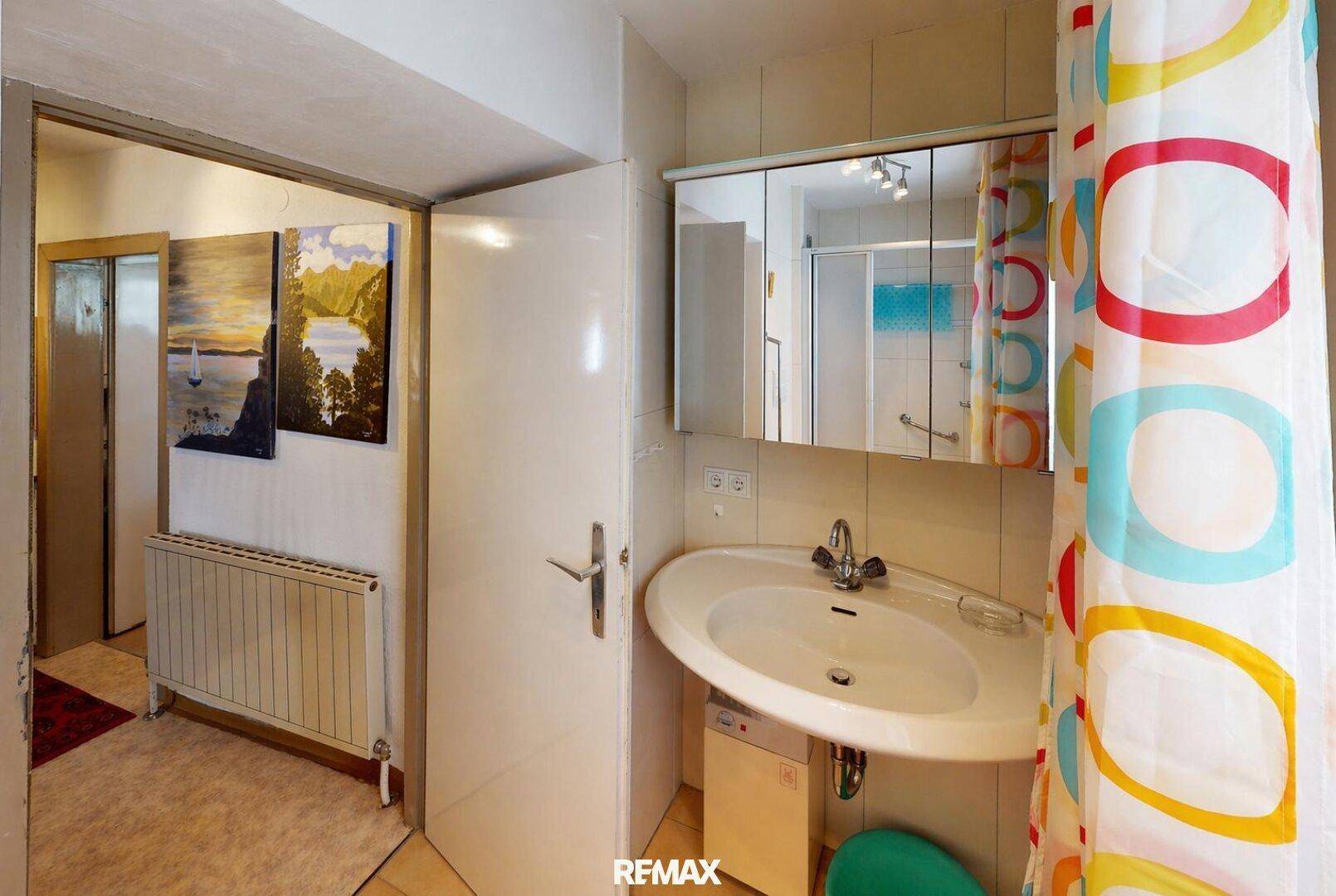 Badezimmer mit Waschbecken, Spiegelschrank und einem farbenfrohen Duschvorhang, angrenzend an einen Flur.