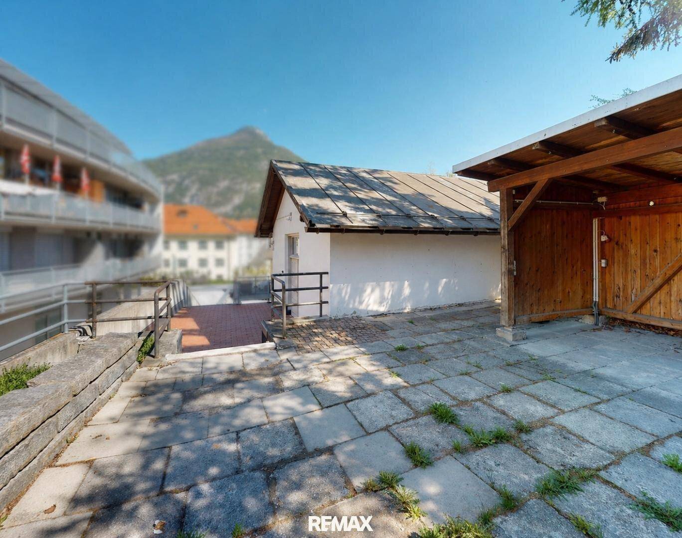 Außenbereich mit gepflastertem Hof, einem kleinen Gebäude und einem Carport im Vordergrund.