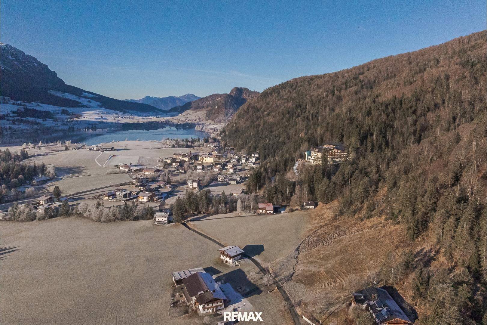 Luftaufnahme einer verschneiten Landschaft mit einem Dorf, einem See und einem großen Gebäude am Berghang.