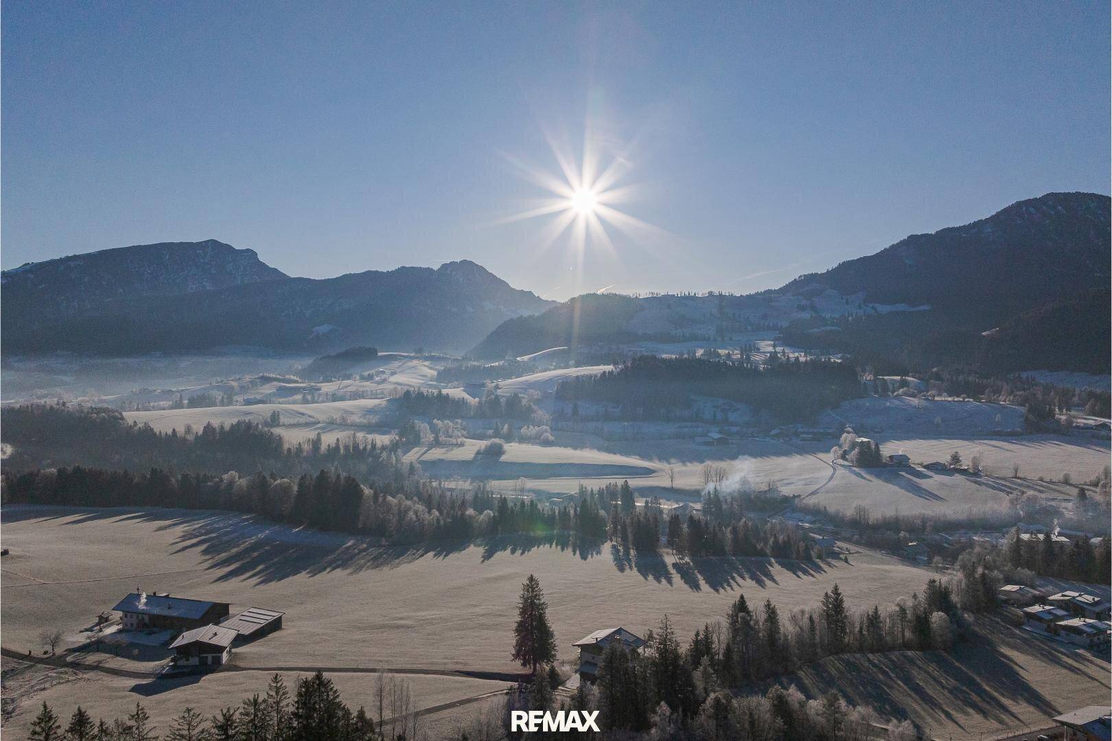 Weitläufige Luftaufnahme einer verschneiten Winterlandschaft mit strahlendem Sonnenschein und Bergen im Hintergrund.