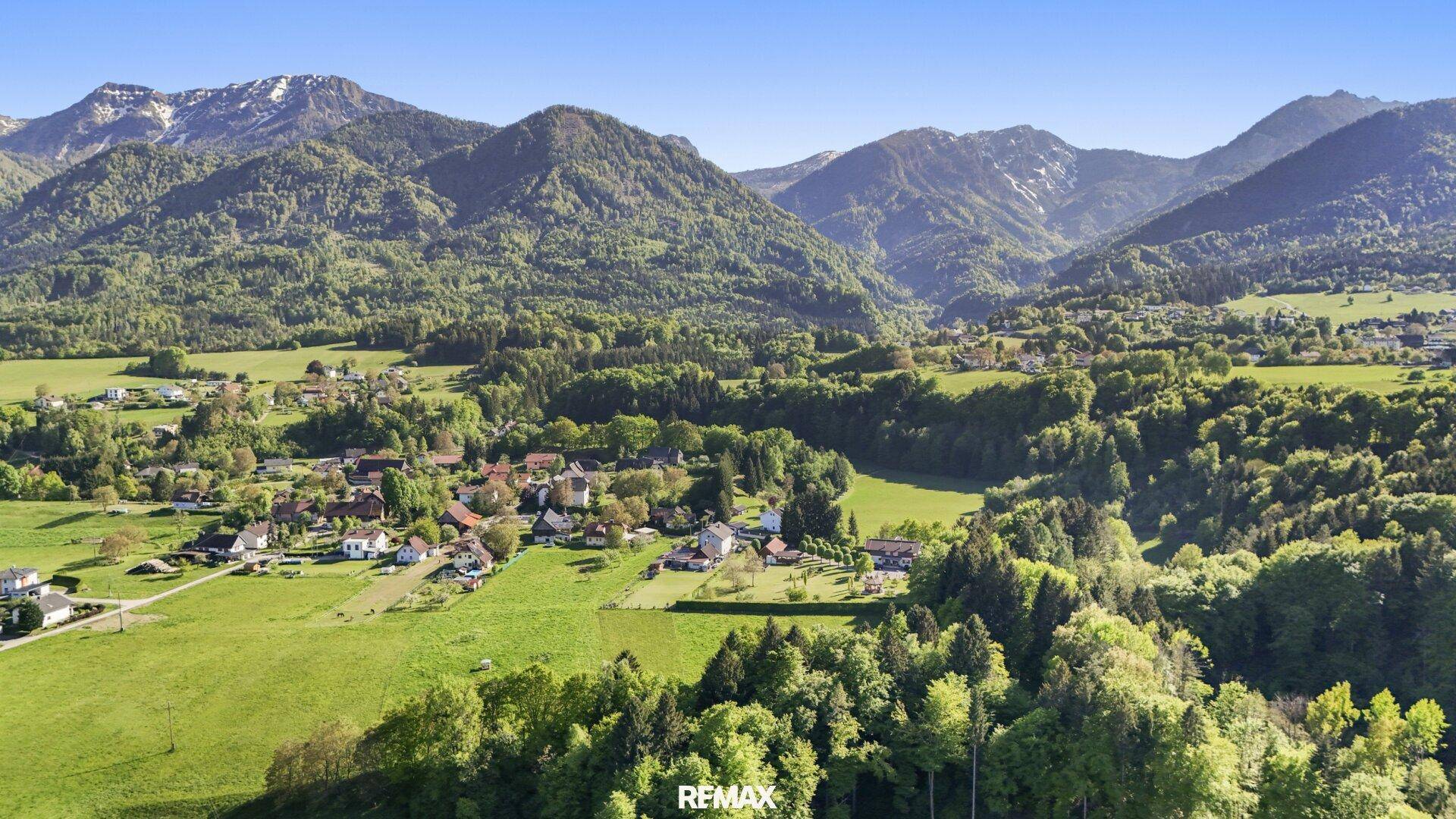 Malerische Landschaft mit grünen Wiesen, Wäldern und einem Dorf vor einer Bergkulisse.