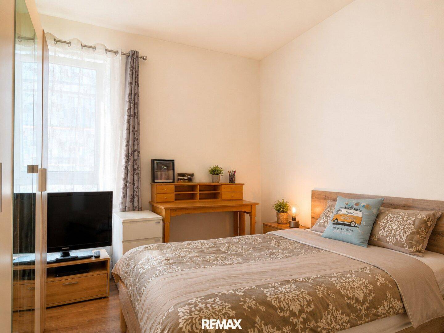 Helles Schlafzimmer mit Holzbett, Schreibtisch und Fenster mit Vorhängen.