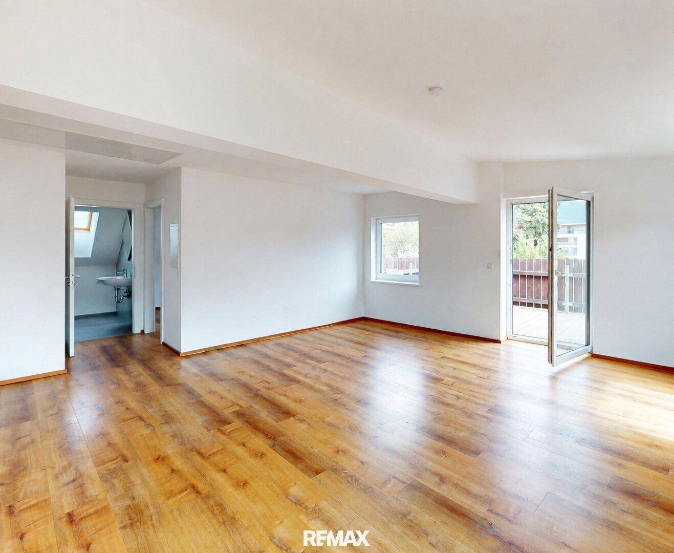 Heller, leerer Wohnbereich mit Holzboden, Fenster und Zugang zu einem Außenbereich.