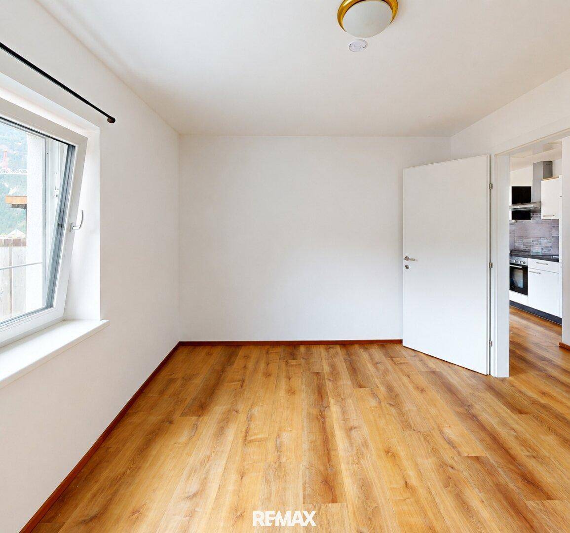 Helles Zimmer mit Holzboden und Fenster, mit Blick auf den Küchenbereich.