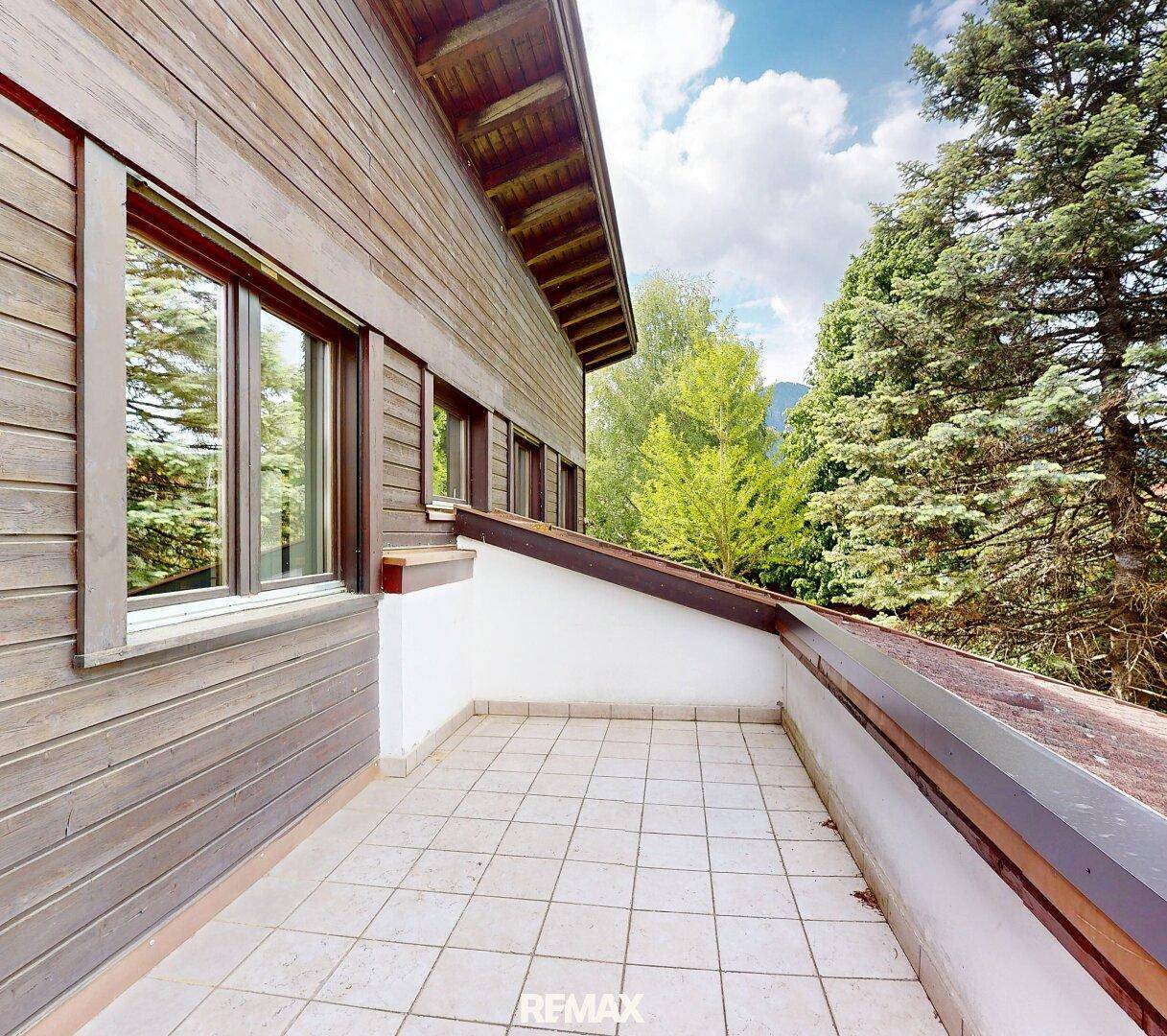 Heller Balkon mit Fliesenboden und Holzwänden, umgeben von grünen Bäumen.