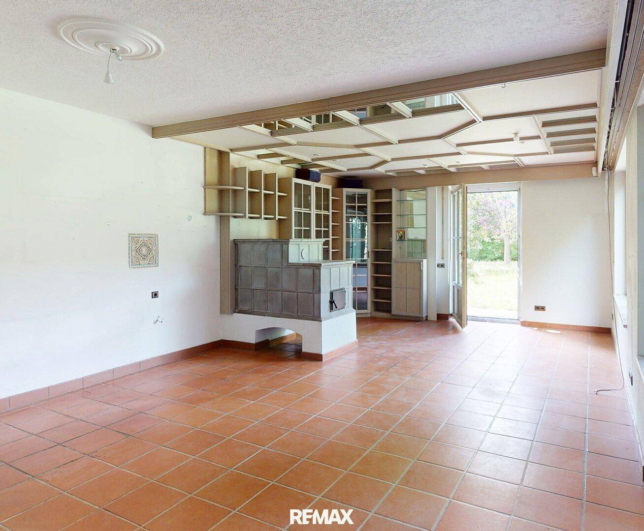 Heller Wohnraum mit Kachelofen, Fliesenboden und Zugang zum Garten oder Balkon.
