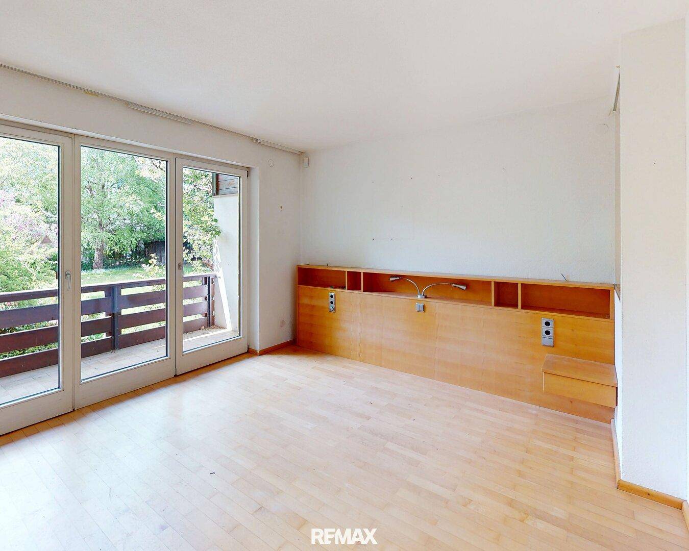 Helles Schlafzimmer mit Holzboden, integriertem Kopfteil und direktem Zugang zum Balkon mit Grünblick.