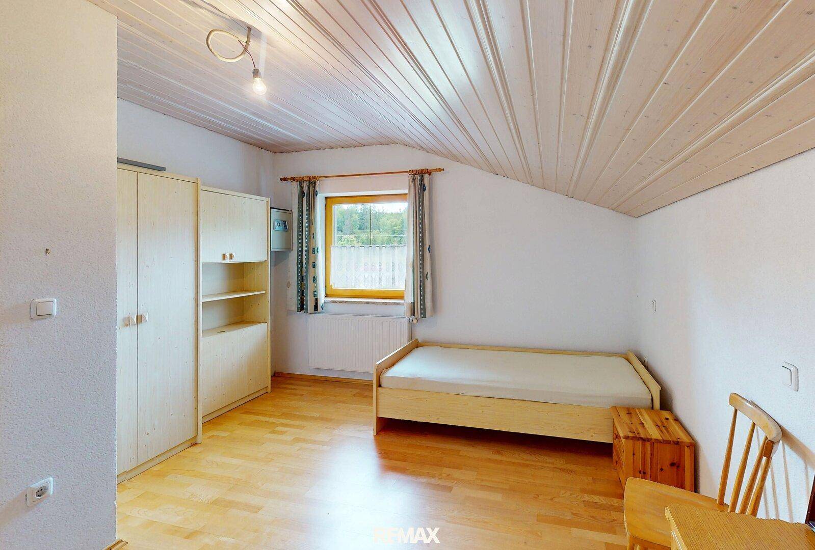 Helles Schlafzimmer mit Holzdecke, Einzelbett und Kleiderschrank, mit Blick ins Grüne.