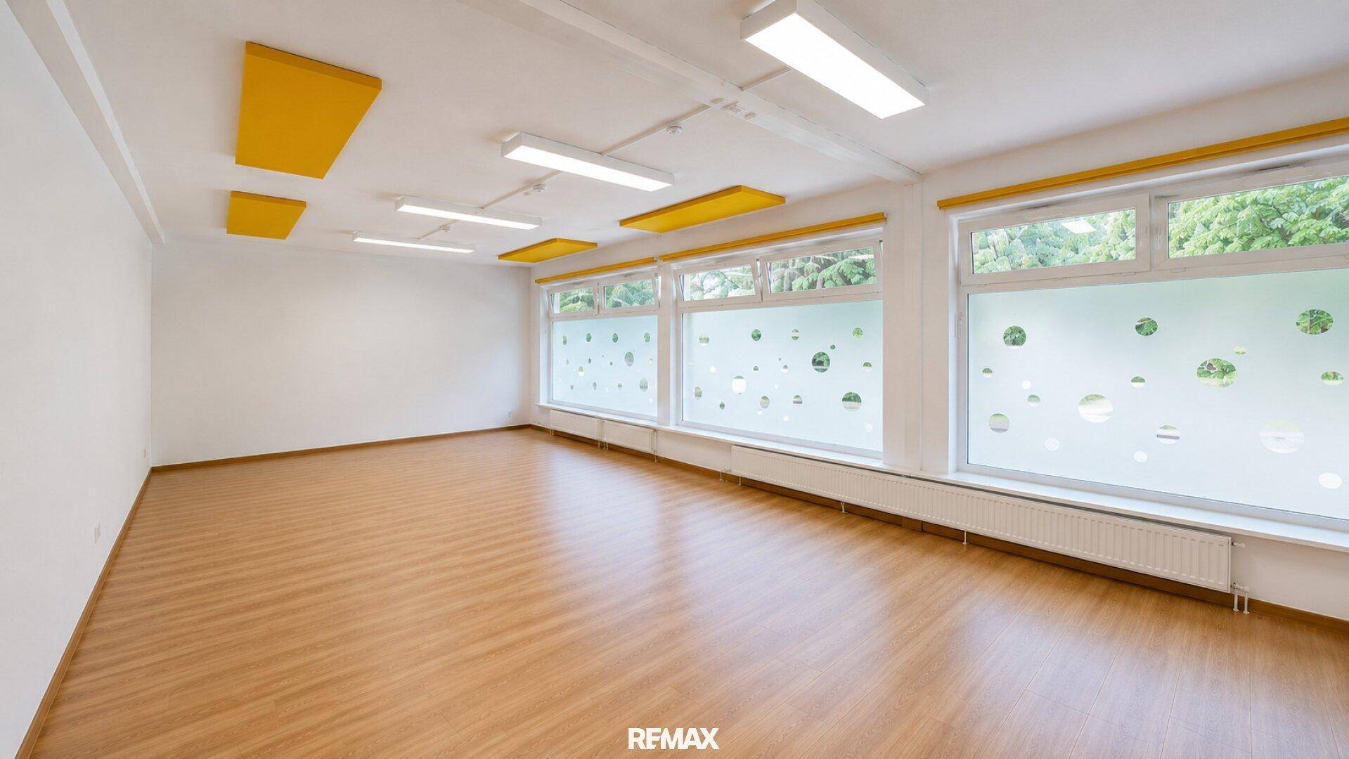 Heller Raum mit Laminatboden, gelben Akustikpaneelen an der Decke und großen Fenstern mit Sichtschutz.