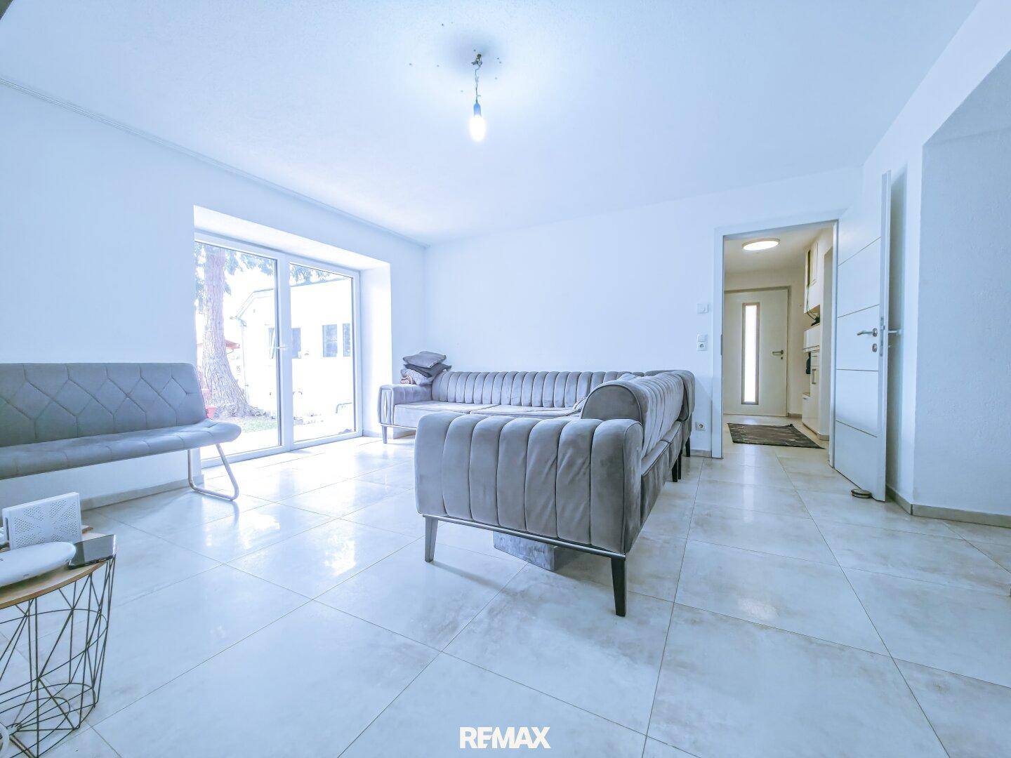 Helles Wohnzimmer mit grauen Sofas, Fliesenboden und großer Glasschiebetür zum Außenbereich.