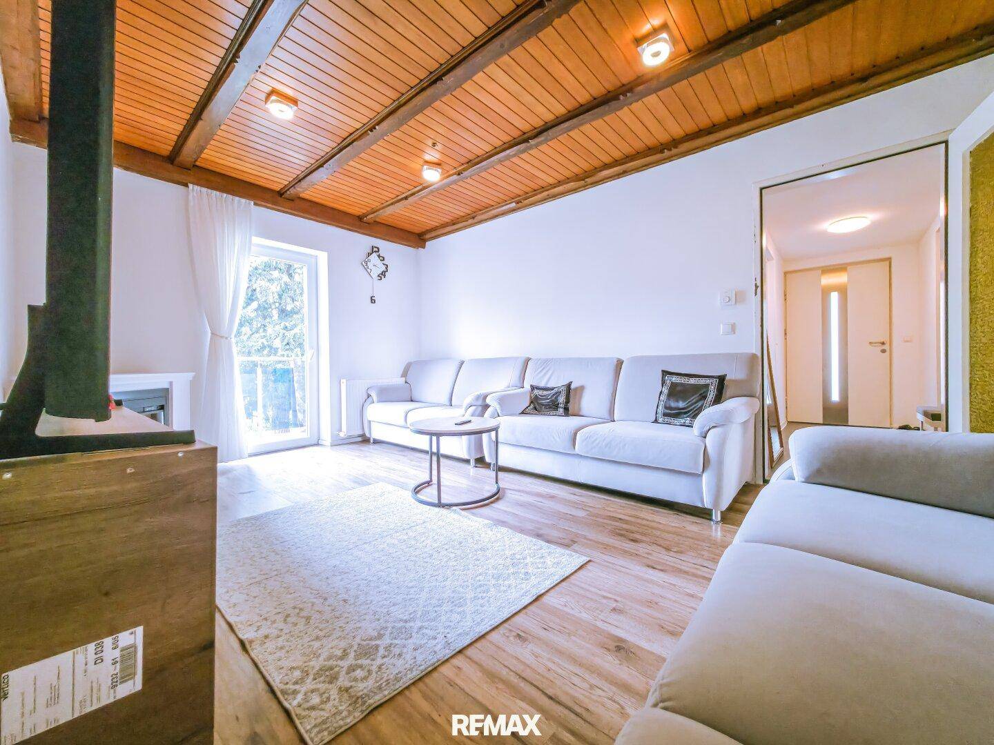 Gemütliches Wohnzimmer mit Holzbalkendecke, hellen Sofas und Zugang zu einem Außenbereich.