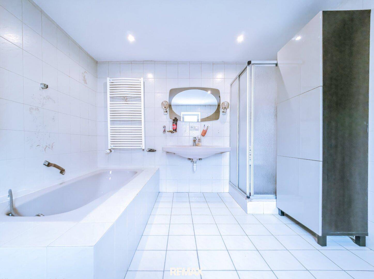 Helles Badezimmer mit weißen Fliesen, Badewanne, Dusche und beheiztem Handtuchhalter.