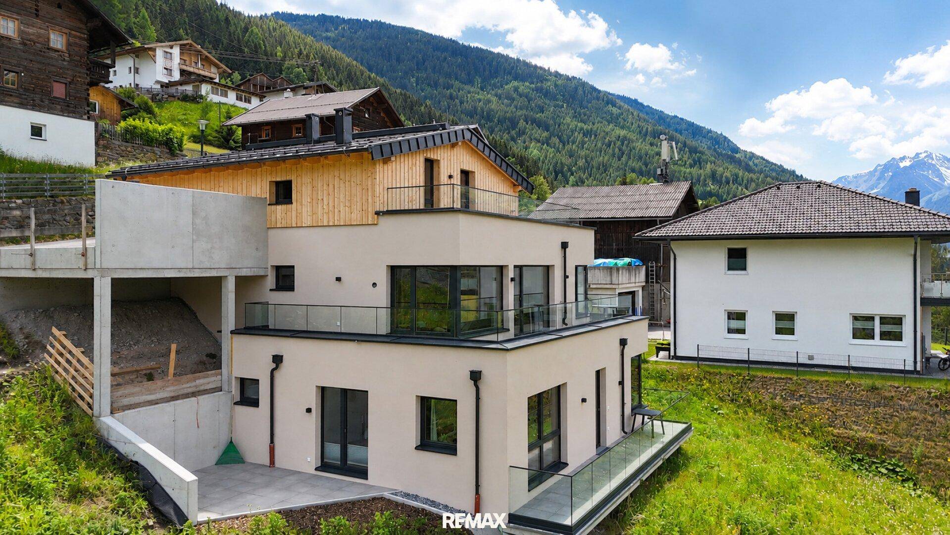 Modernes Haus mit Flachdach und Balkonen in alpiner Umgebung, umgeben von grünen Hügeln und traditionellen Gebäuden.