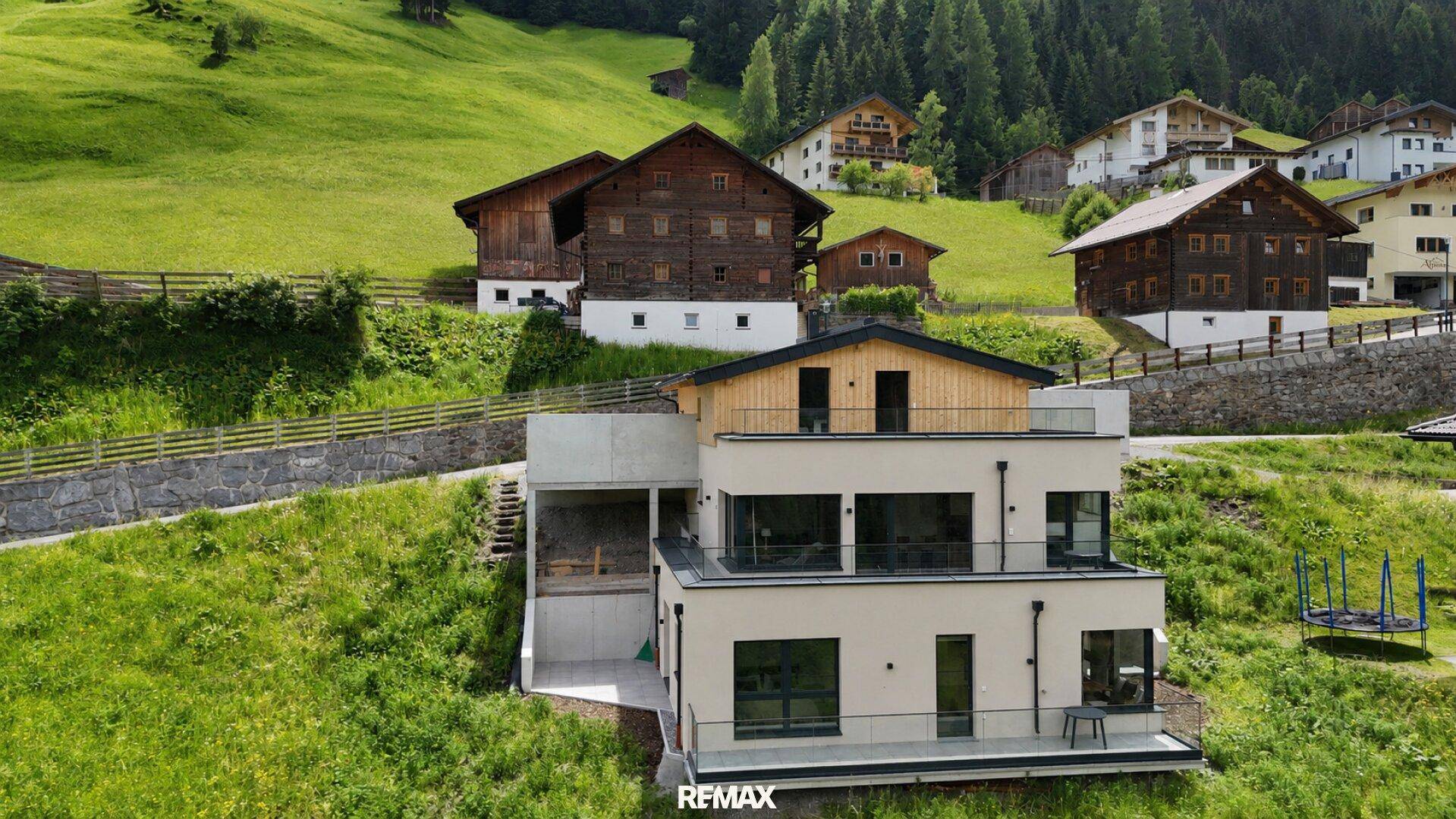 Modernes Gebäude in alpiner Landschaft mit grünen Hängen und traditionellen Holzhäusern im Hintergrund.