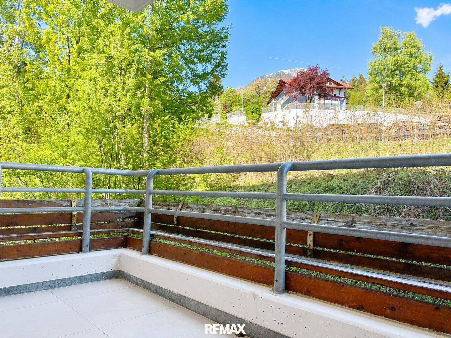 Geräumiger Balkon mit Metallgeländer und Blick ins Grüne sowie auf ein entferntes Haus.