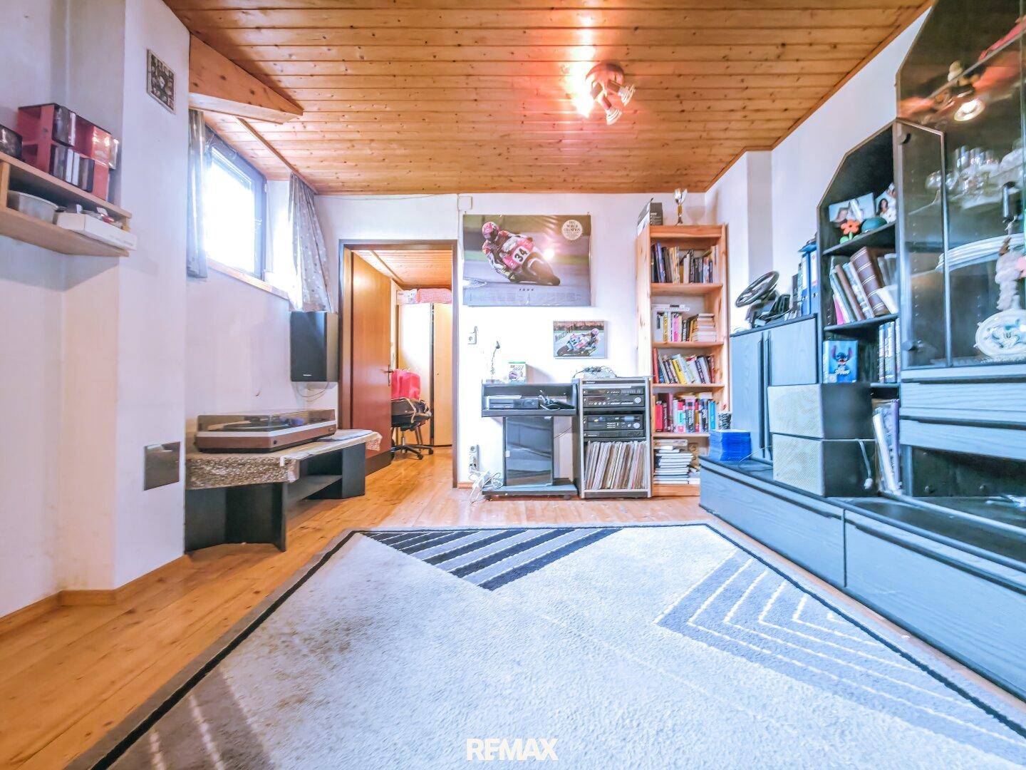 Gemütliches Wohnzimmer mit Holzdecke, Parkettboden und einem Teppich, eingerichtet mit Büchern und Unterhaltungselektronik.