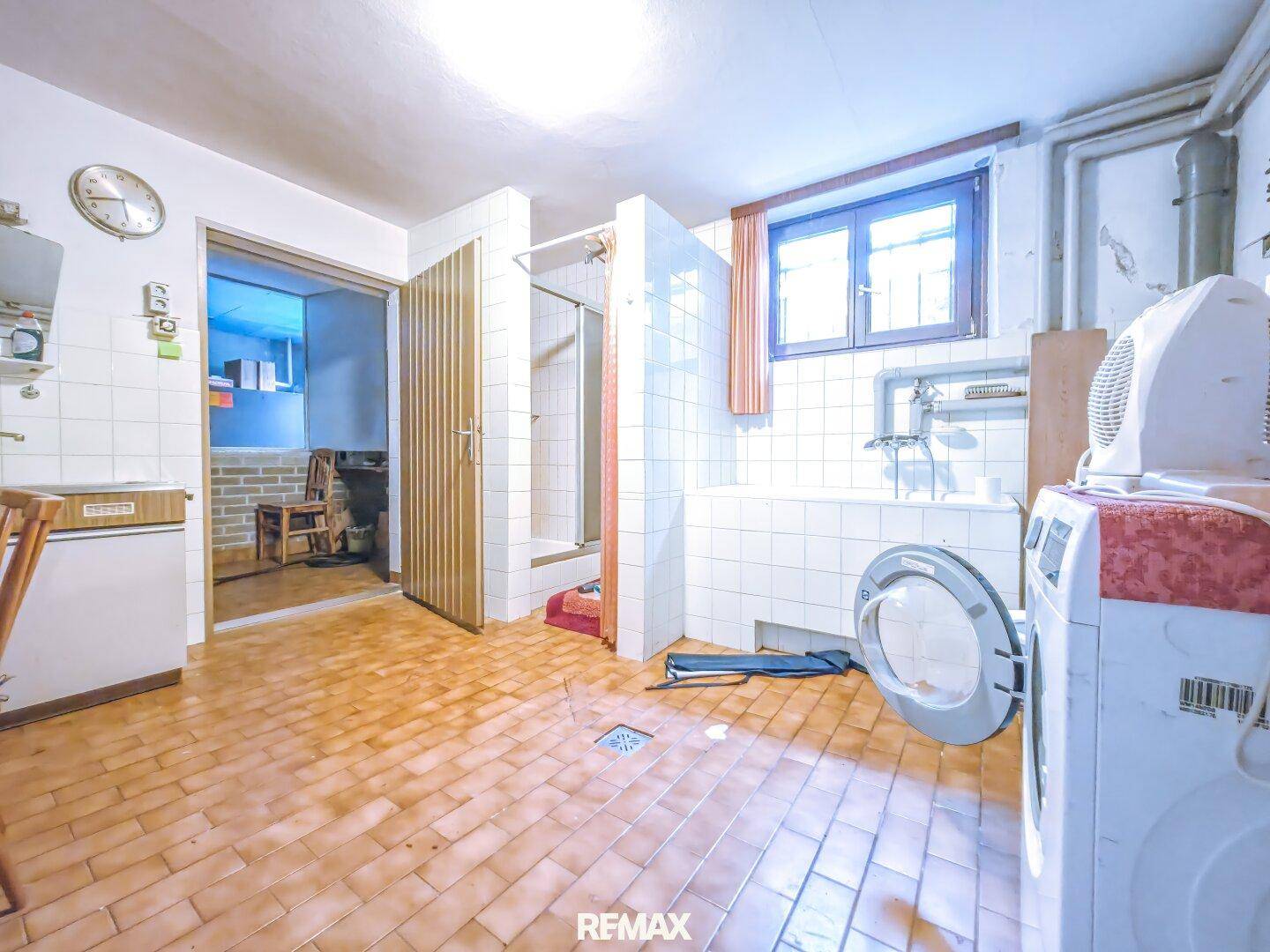 Funktionales Badezimmer mit Fliesenboden, Badewanne, Dusche und Waschmaschine, beleuchtet durch ein Fenster.