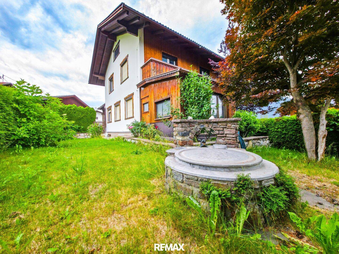 Gartenbereich mit Rasenfläche, einem dekorativen Brunnen und dem Haus im Hintergrund, umgeben von Bäumen.