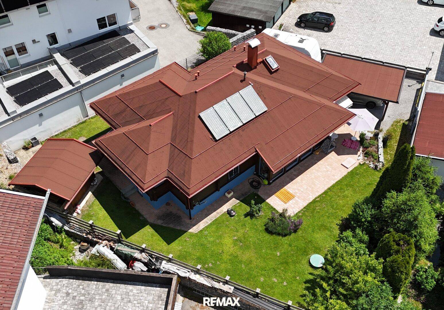 Luftaufnahme des Holzhauses mit Solarpaneelen auf dem roten Dach und umliegenden Gebäuden.