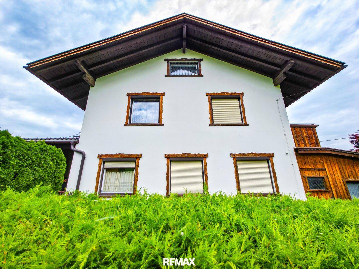 Vorderansicht des Hauses mit weißer Fassade, Holzdetails und Rollläden an den Fenstern.