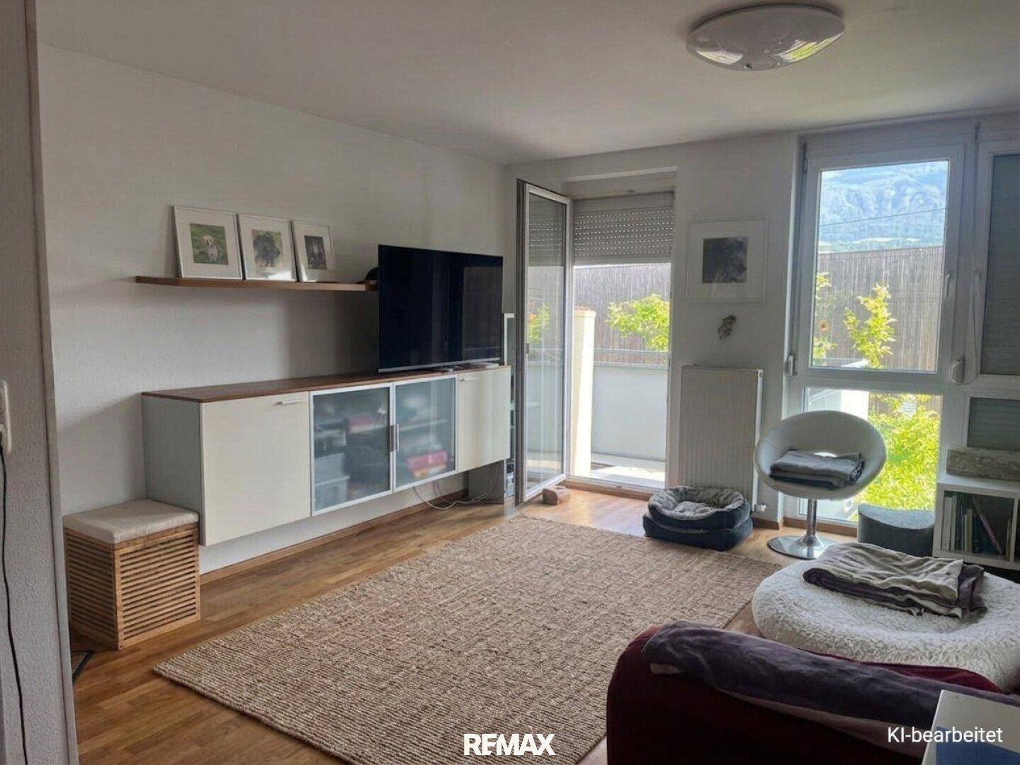 Gemütlicher Wohnbereich mit Fernseher, Holzboden und viel Tageslicht durch große Fenster.