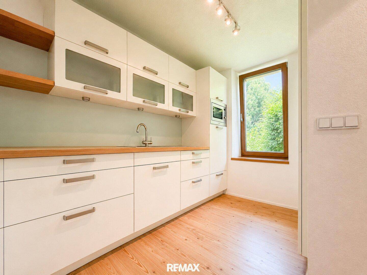 Funktionale Küche mit weißen Schränken, Holz-Arbeitsplatte und Fenster mit Grünblick.