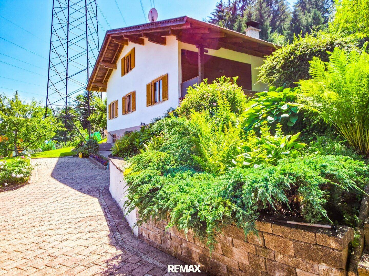 Haus mit gepflasterter Zufahrt und üppiger Bepflanzung, im Hintergrund ein Strommast.