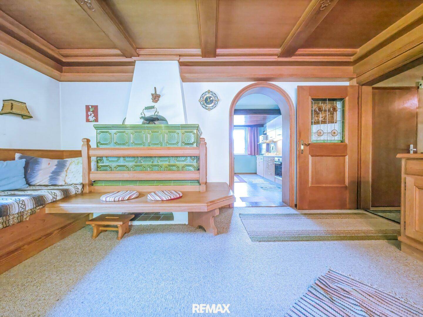 Wohnzimmer mit traditionellem Kachelofen, Holzbänken und einem Durchgang zu weiteren Räumen.