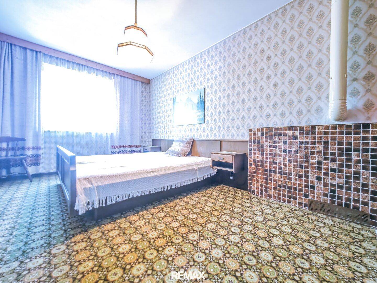 Schlafzimmer mit gemusterter Tapete, Bett und einem gefliesten Heizelement.
