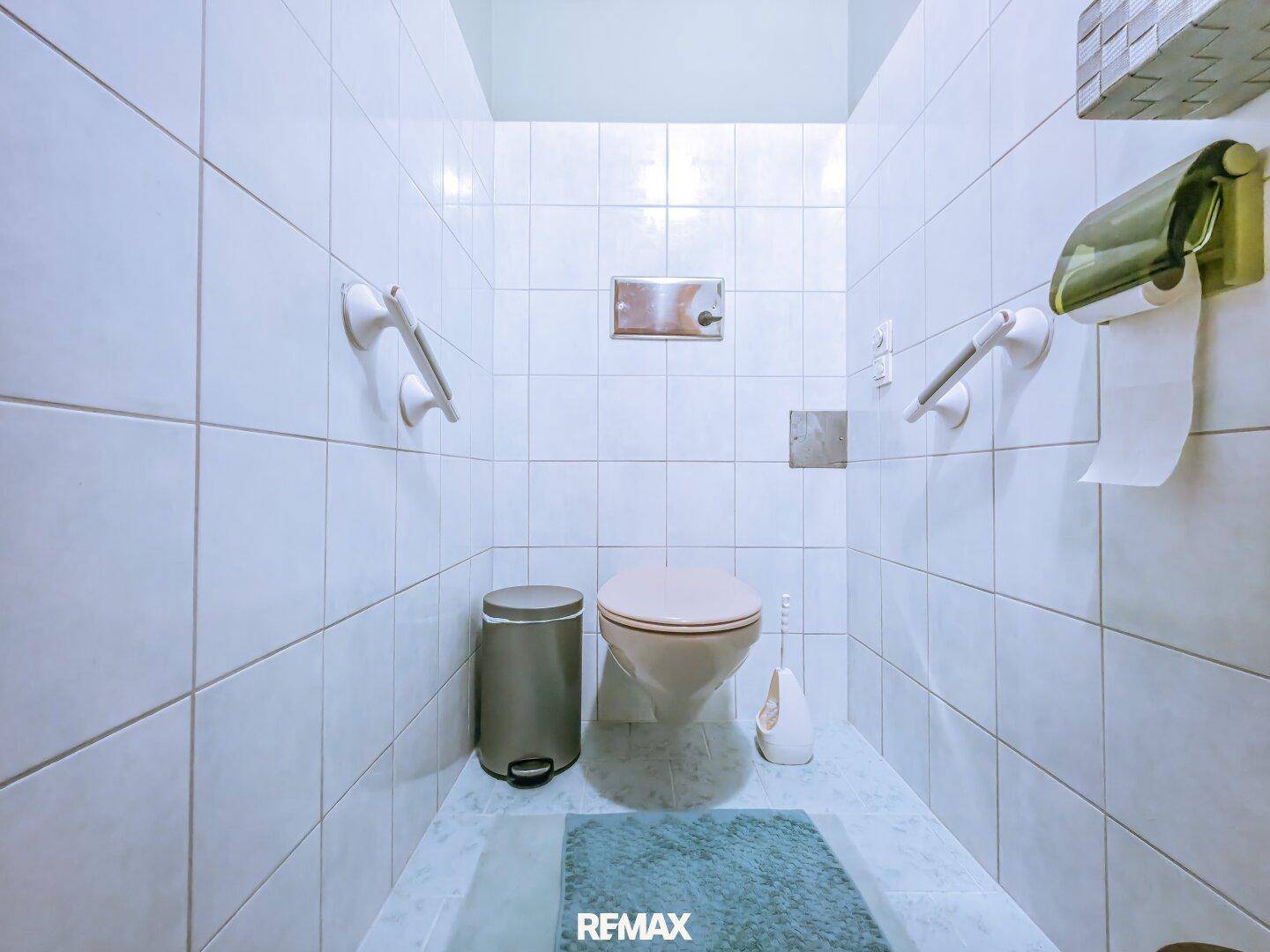 Separates WC mit weißen Fliesen, Toilette und Haltegriffen für zusätzliche Sicherheit.