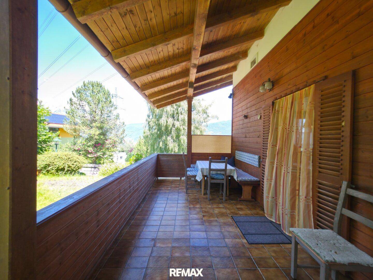 Überdachter Balkon mit Holzbänken, gefliestem Boden und schöner Aussicht ins Grüne.