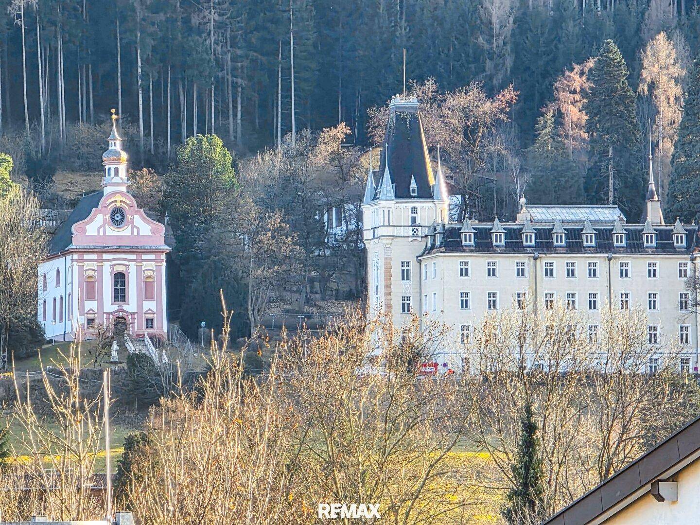 Historisches Gebäude und Kirche vor einem bewaldeten Berghang.