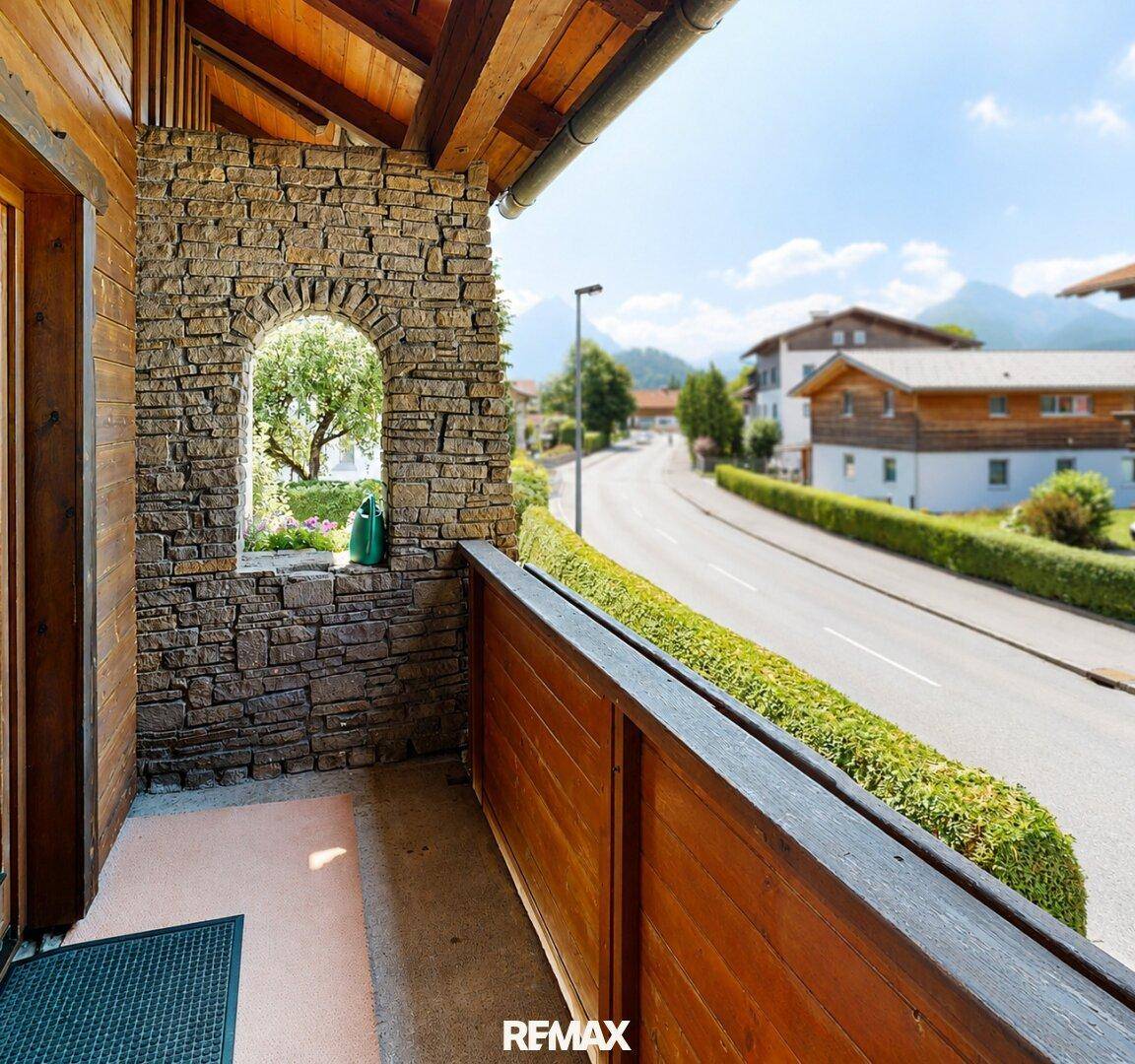 Gemütlicher Balkon mit Steinmauer, Holzgeländer und Blick auf die Straße und Berge.