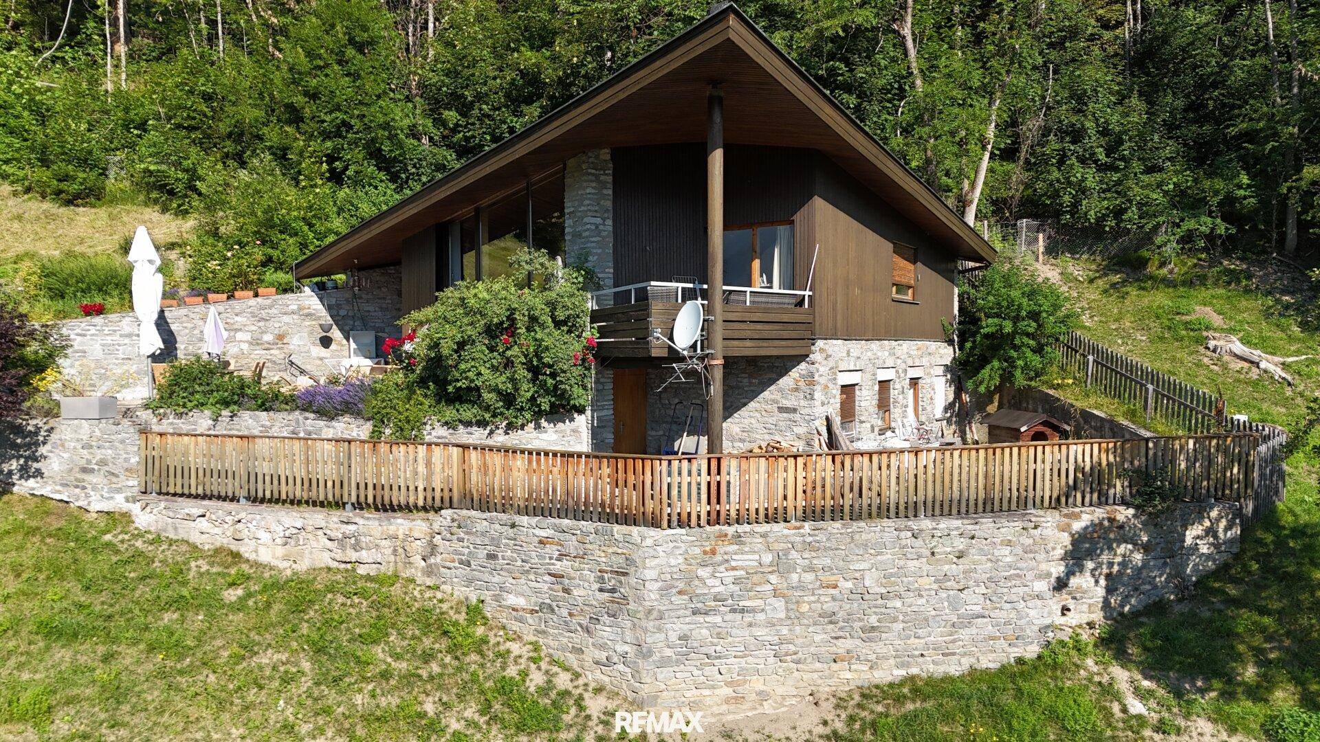 Charmantes Haus mit Steinfassade und Holzverkleidung, eingebettet in eine grüne Hanglage mit üppiger Vegetation.