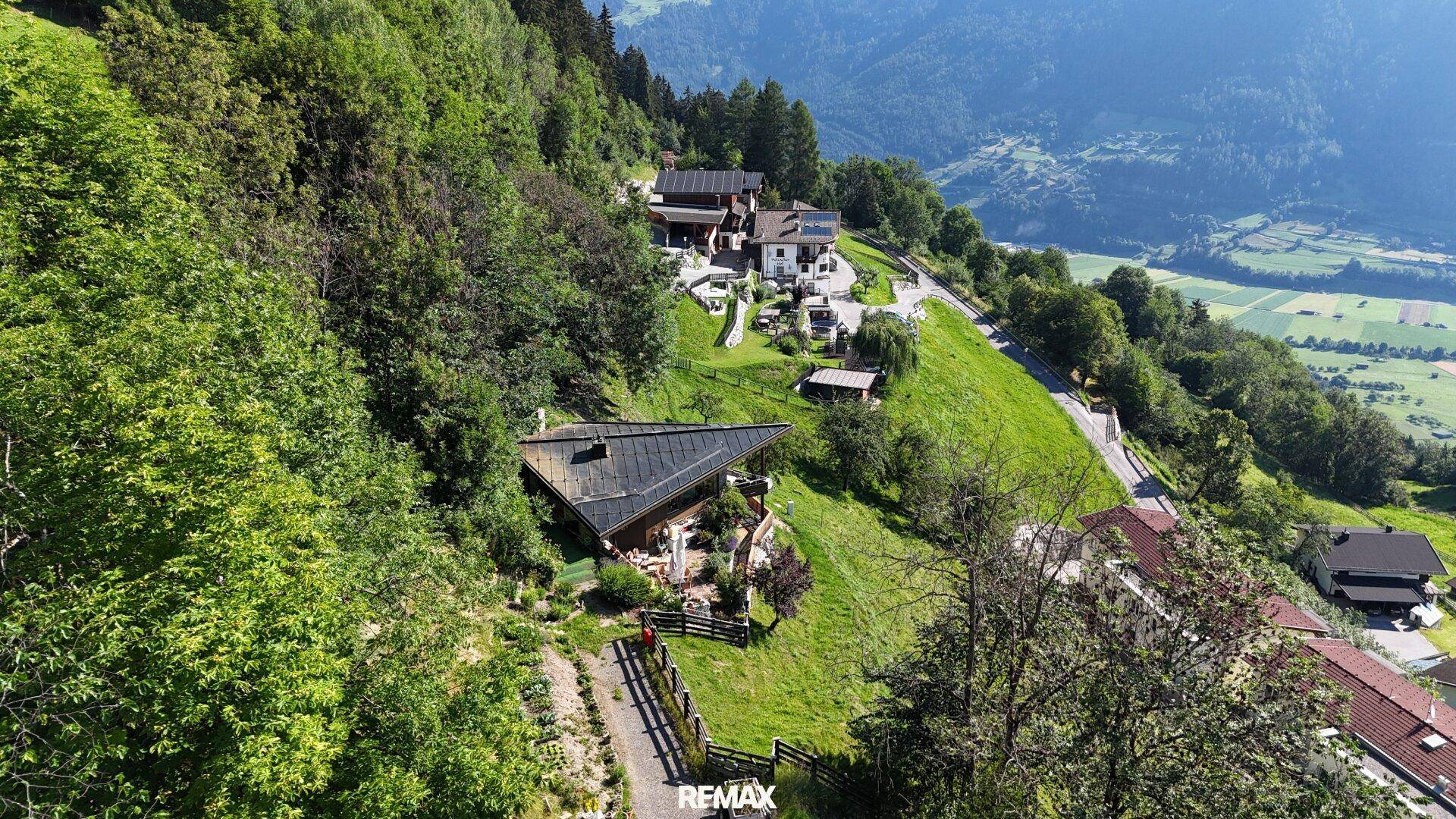 Luftaufnahme des Anwesens auf einem grünen Hang, umgeben von Wald und weiteren Häusern.