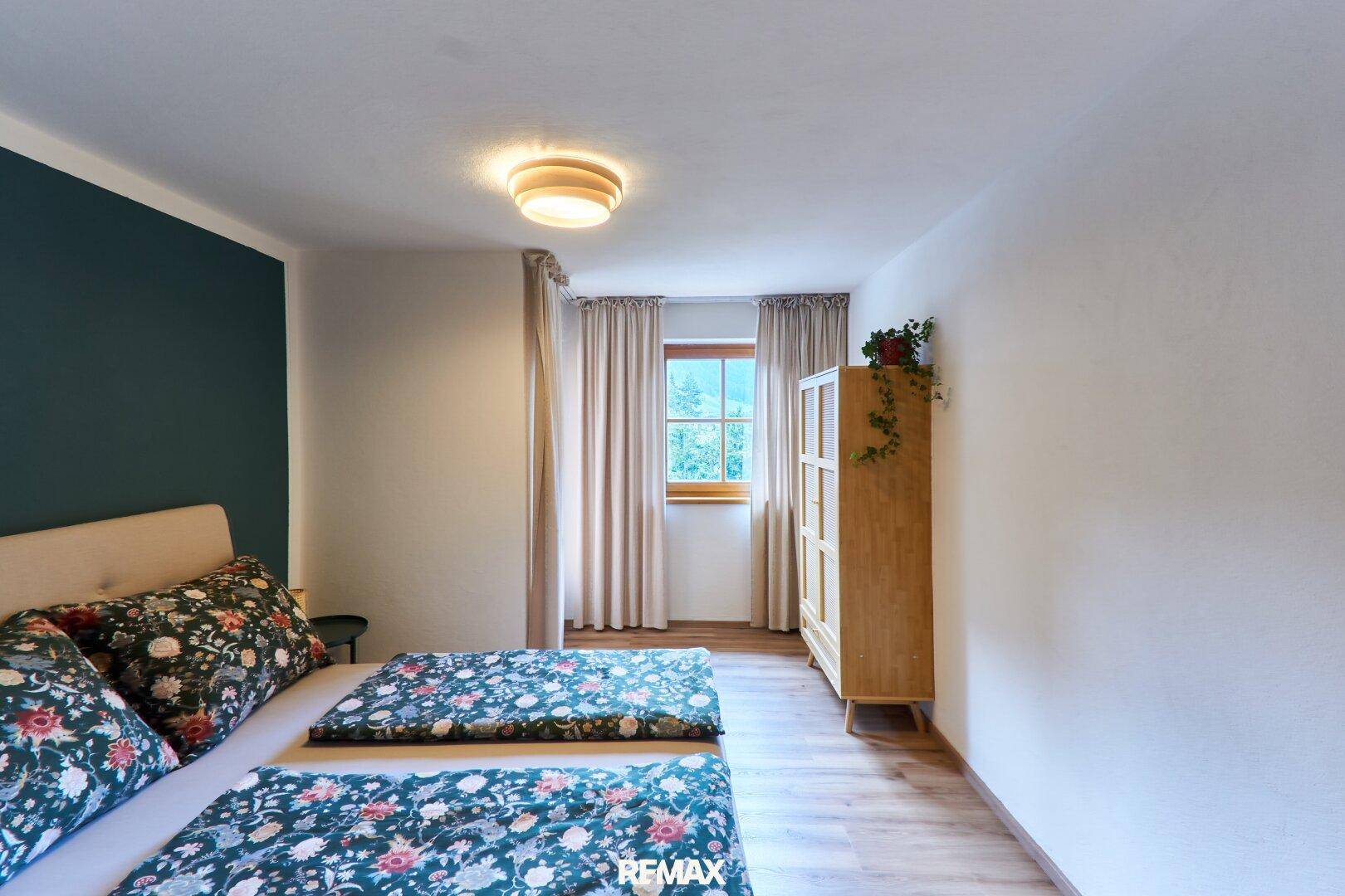 Helles Schlafzimmer mit Holzboden, floraler Bettwäsche und einem Fenster mit Grünblick.