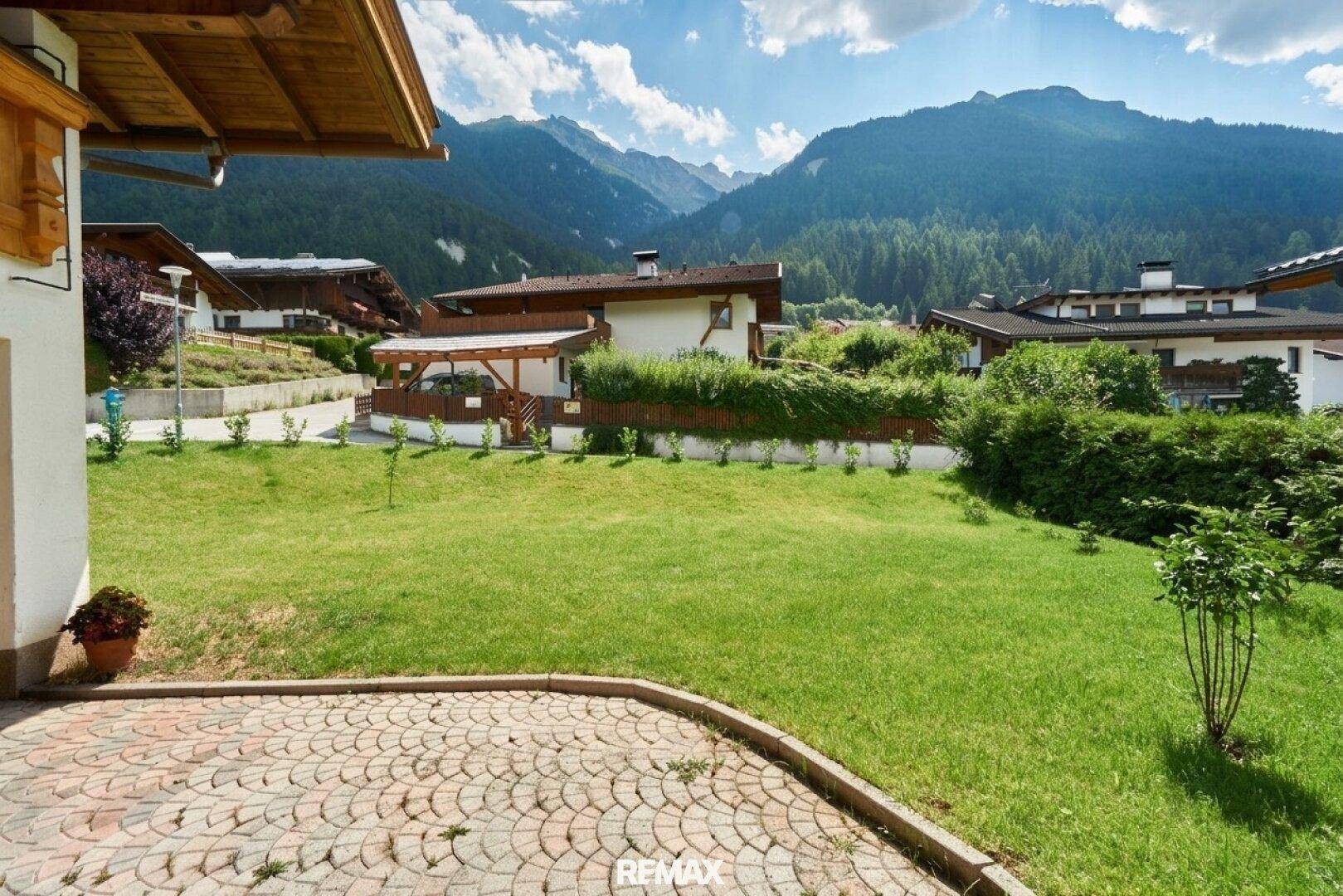 Gepflegter Gartenbereich mit Rasenfläche und gepflasterter Terrasse vor Bergkulisse.