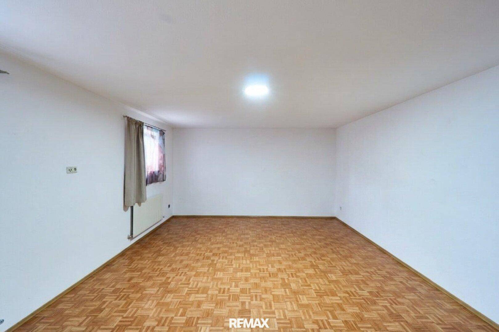 Leerer Raum mit Parkettboden und einem Fenster, das Tageslicht hereinlässt.