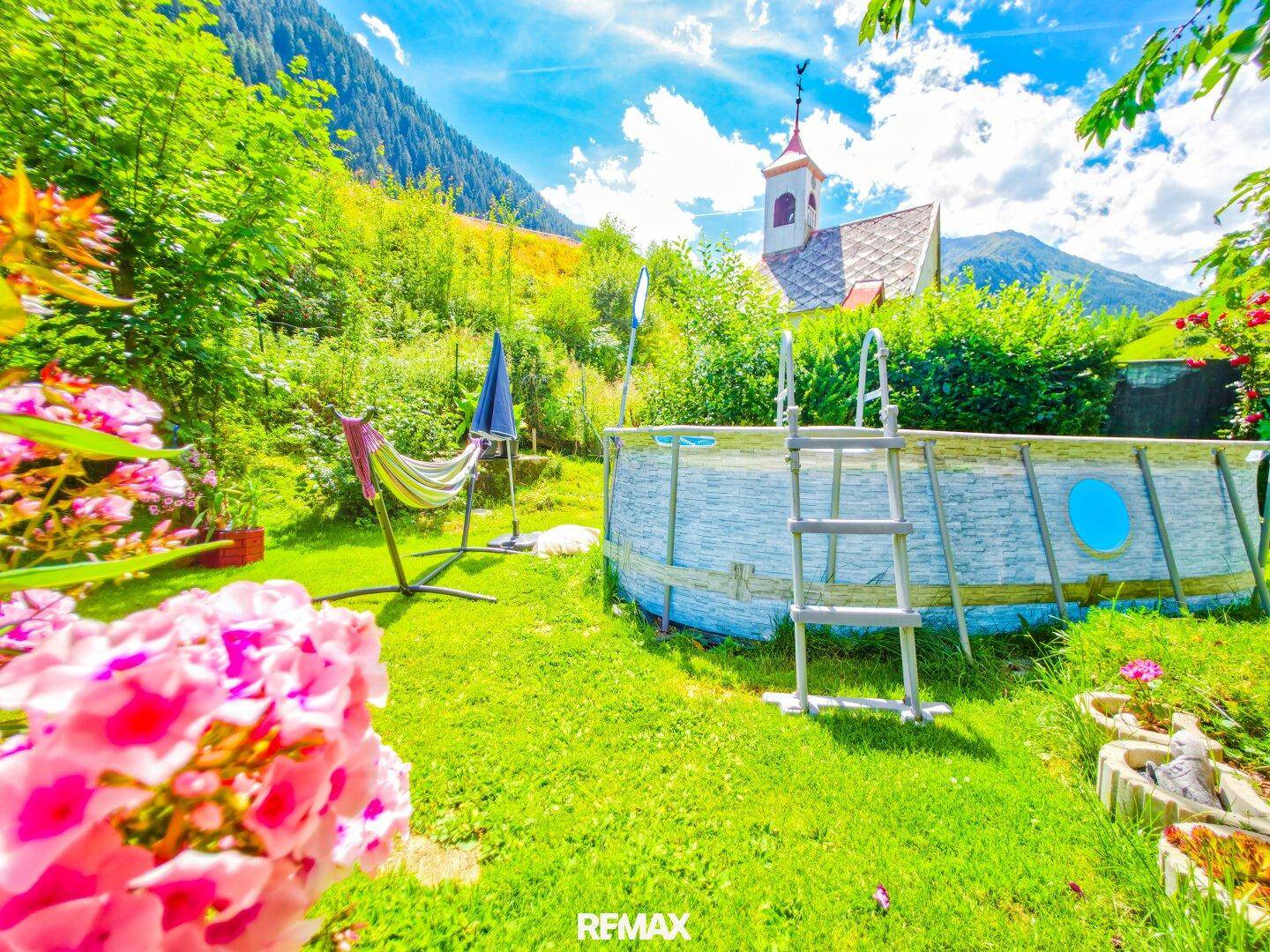 Sonniger Garten mit Aufstellpool und Hängematte, mit Blick auf eine kleine Kapelle und Berge.