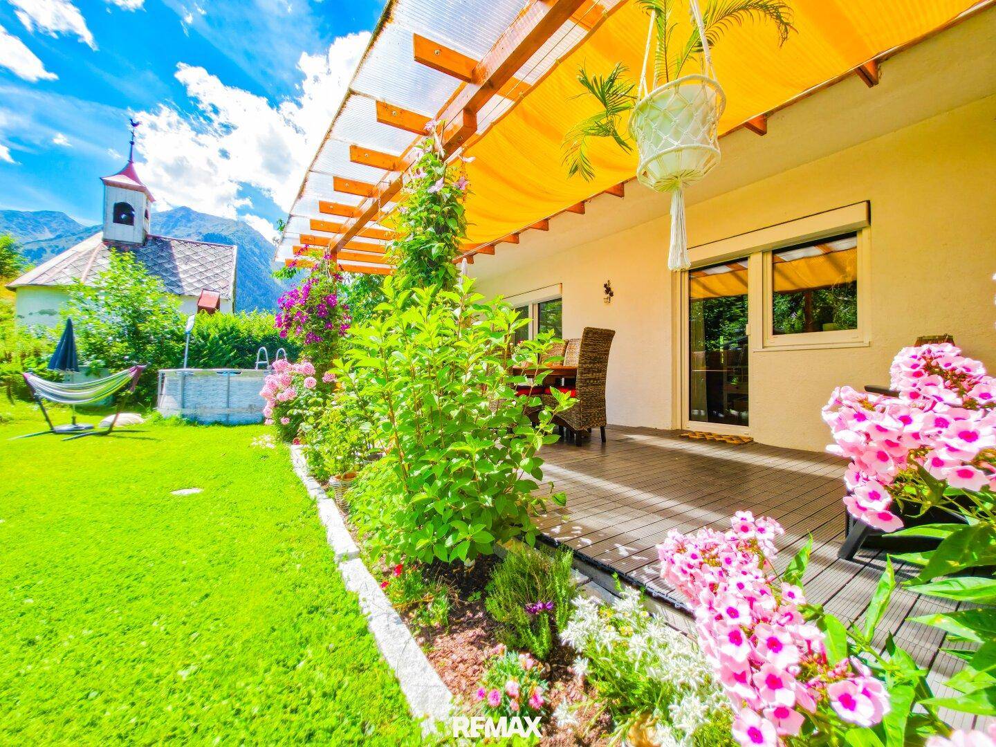 Gemütliche Terrasse mit Pergola und blühenden Blumenbeeten, angrenzend an einen sonnigen Garten.